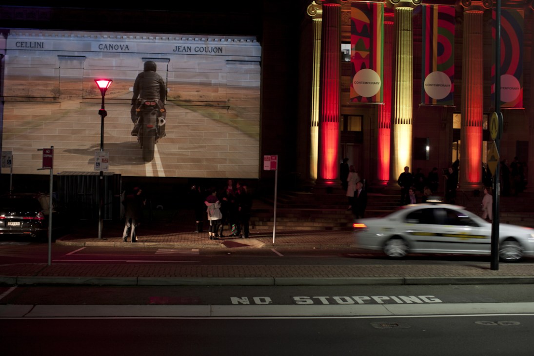 Night exterior of the John Kaldor Family Gallery with red-lit classical columns and a large video screen displaying contemporary art.