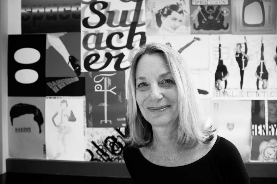 Paula Scher, principal at Pentagram, posed in front of a wall displaying her graphic design work in black and white.