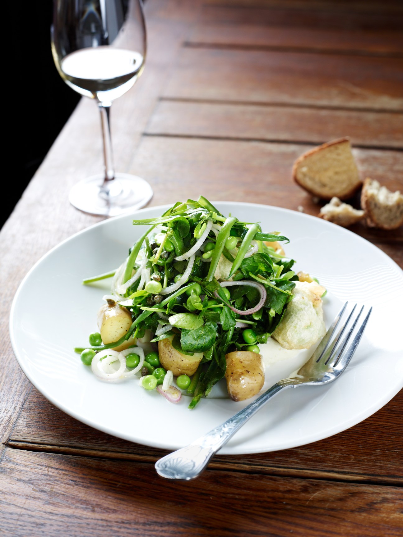 Pea fritter salad with goats curd, watercress, pea shoots, shallots and Jersey Royal potatoes on a white plate with wine glass and bread.