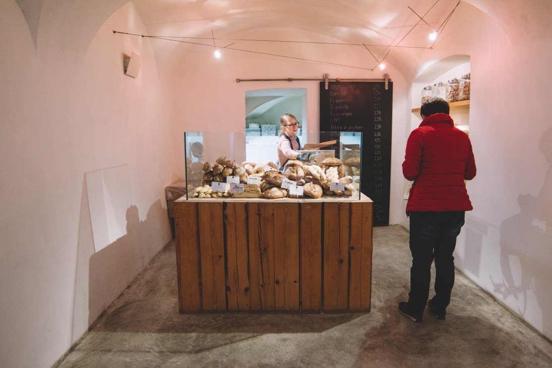 Customer in red jacket at Pekarna Osem bakery counter displaying various fresh breads and baked goods.