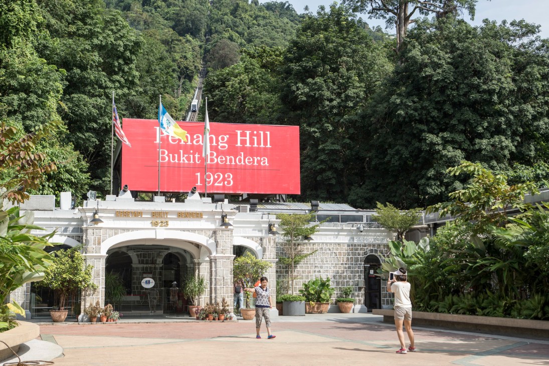 Base station of Penang Hill funicular railway with red sign reading 'Penang Hill Bukit Bendera 1923' and historic stone building.