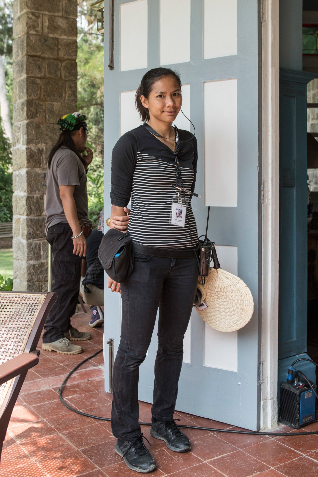 Set PA Krystle Lim stands on a colonial-era veranda holding a woven hat and wearing a production badge.