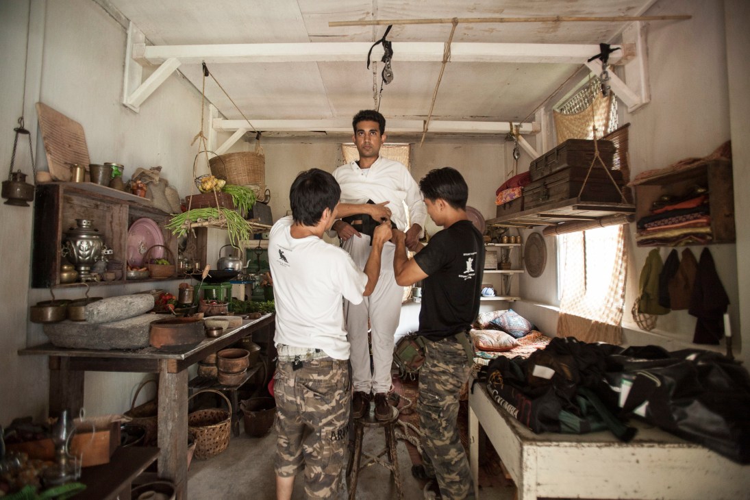 Malaysian actor Ash Nair being prepared for filming in a period room set with two crew members adjusting his costume.