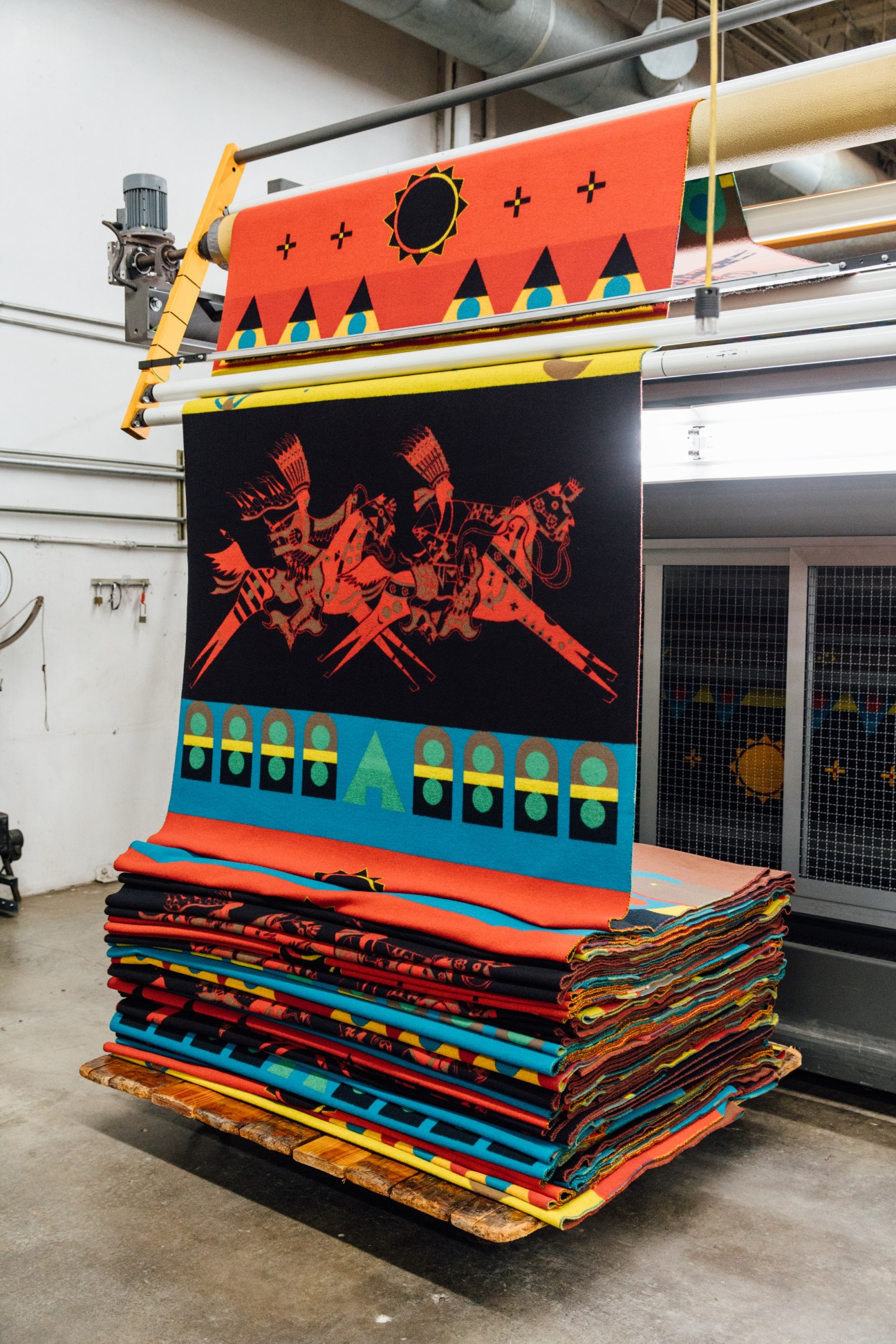 Colorful blankets stacked in a warehouse featuring indigenous geometric patterns and motifs in orange, blue, yellow and black.