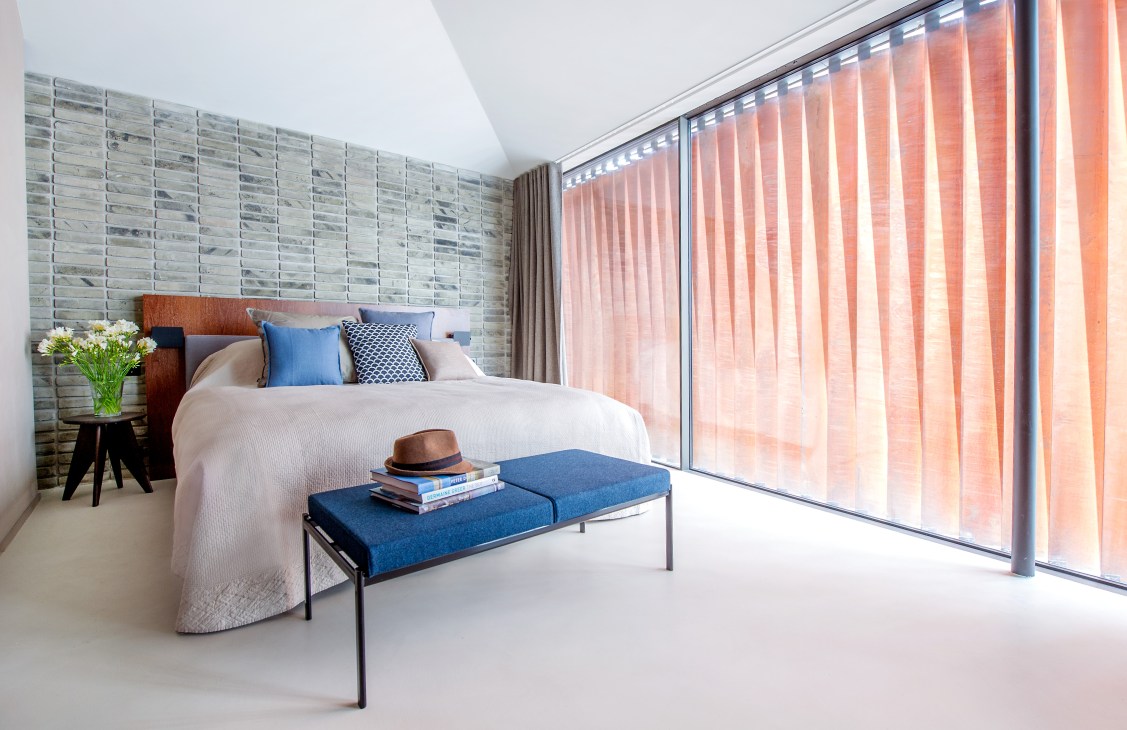 Modern bedroom with brick accent wall, white bed with blue pillows, blue bench, and floor-to-ceiling windows with sheer curtains.