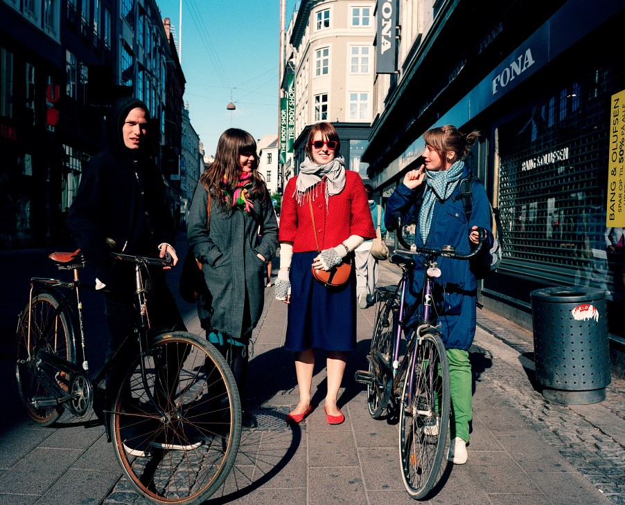 Group of four young people with bicycles on a sunny Copenhagen street lined with shops and buildings.