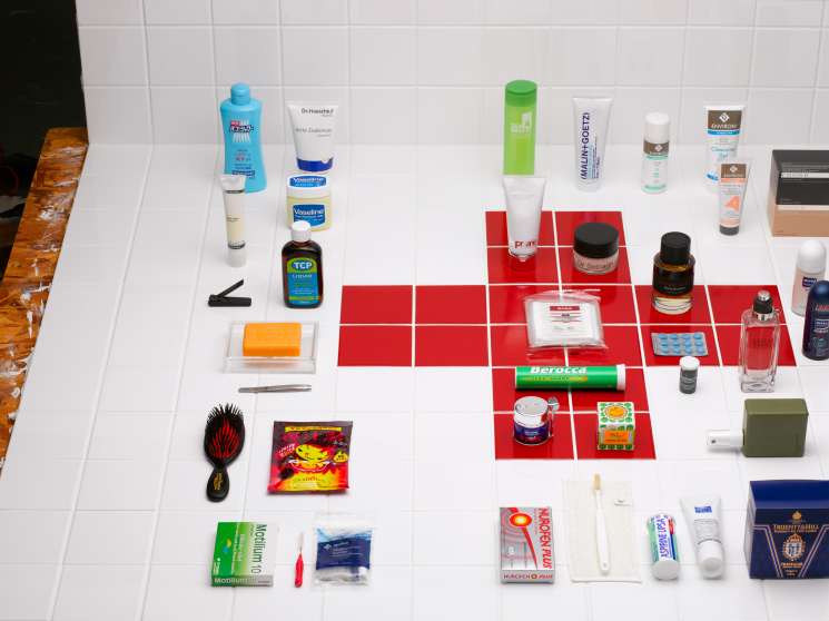 Flat lay of 33 bathroom and personal care products arranged on white tiles with red squares, including cosmetics, medications, and grooming tools.