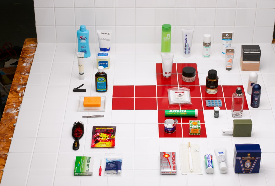 Flat lay of 33 bathroom and personal care products arranged on white tiles with red squares, including cosmetics, medications, and grooming tools.