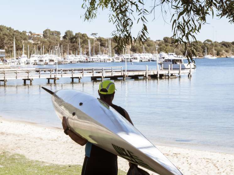 Kayaker in neon helmet carrying boat on Swan River beach at Bicton Baths with moored yachts behind.