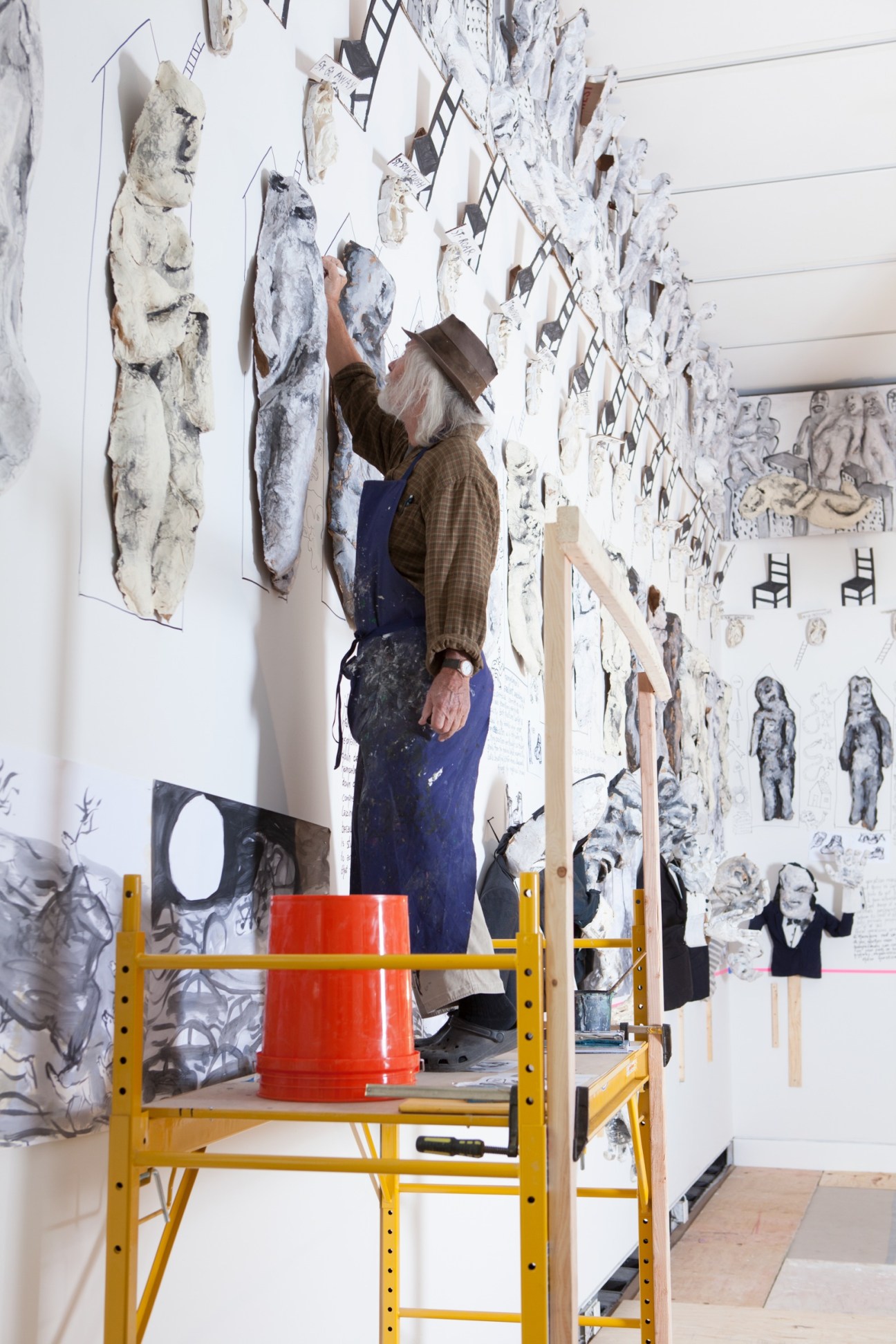 Peter Schumann working on a ladder installing artwork on gallery walls covered with drawings and sketches.