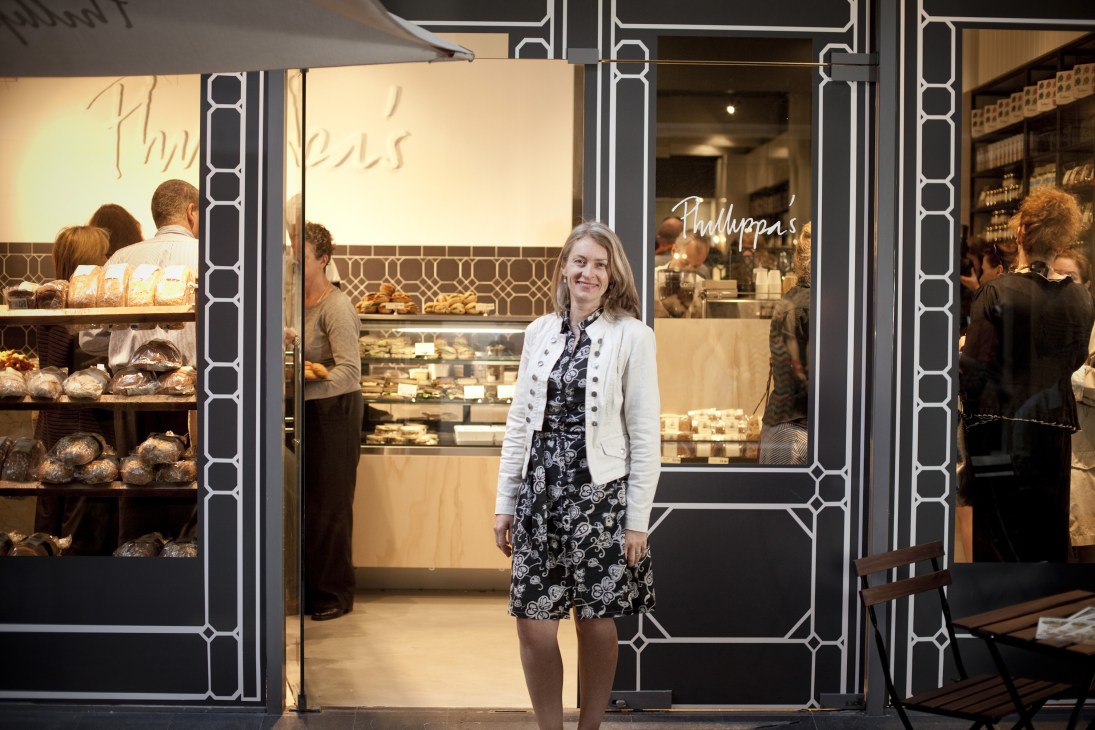 Philippa Grogan stands outside her Melbourne bakery storefront, displaying baked goods and pastries behind glass windows.