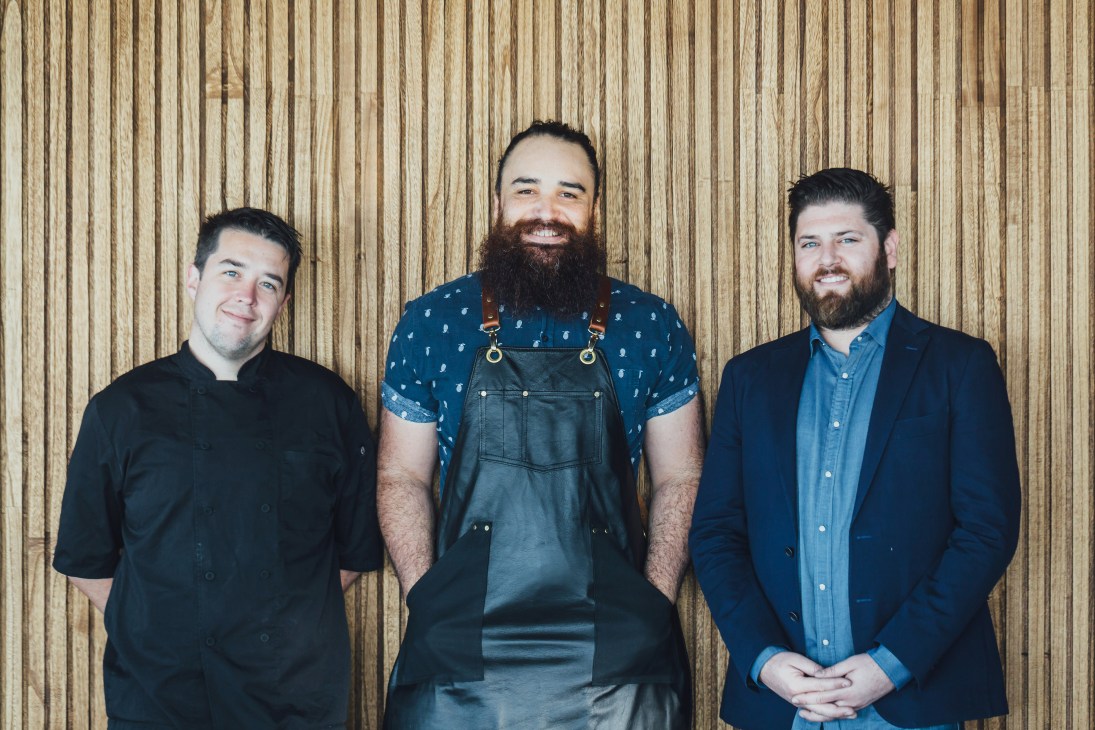 Three men standing against a wooden wall backdrop, with the center man wearing a denim apron.