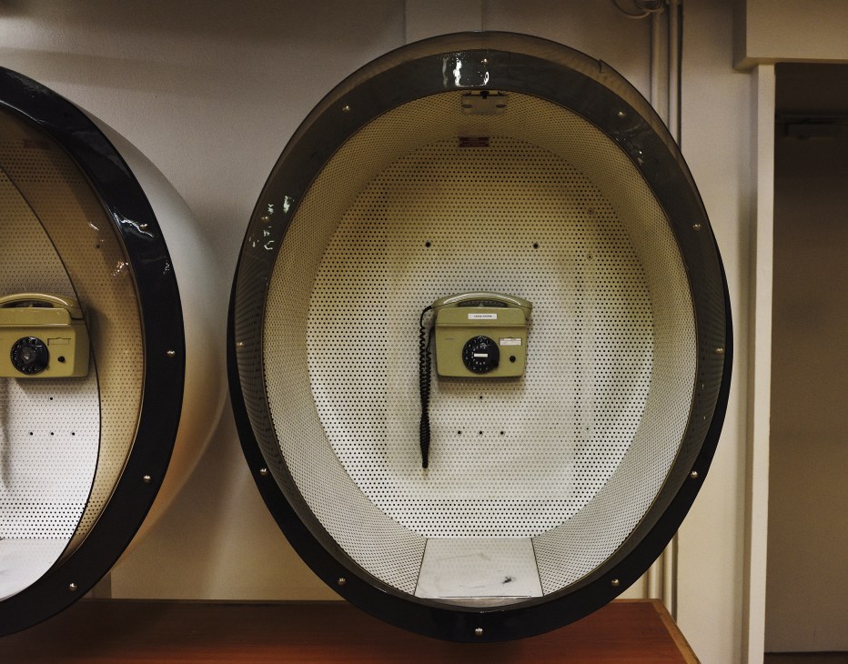 Vintage yellow rotary telephones mounted inside oval-shaped beige perforated panels on a wall.