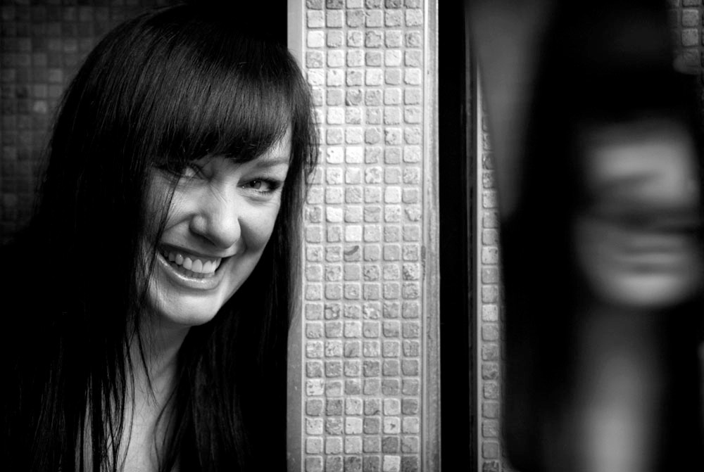 Black and white portrait of Phoebe Greenberg smiling beside a brick wall.