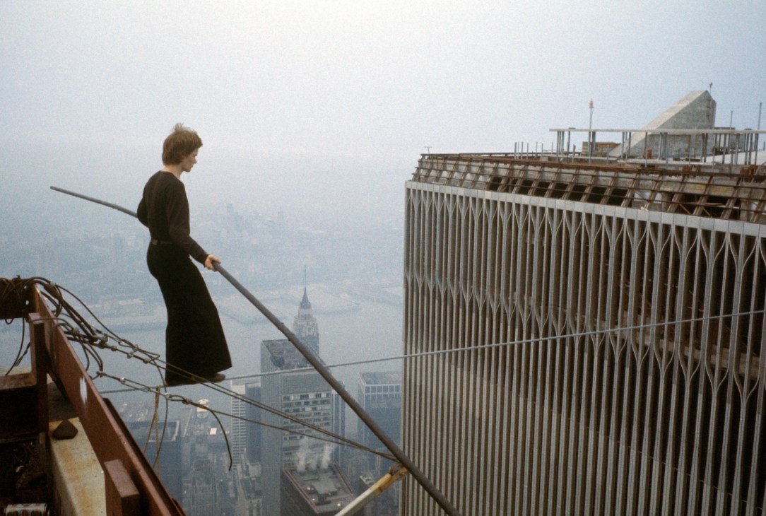 A man in dark clothing holds a long pole while standing on a wire strung between two tall buildings high above a city skyline.