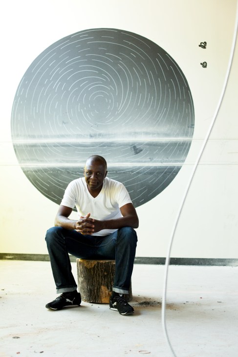 Chef Shola Olunloyo sits on a wooden block against a circular wall art installation at Piazza at Schmidts.