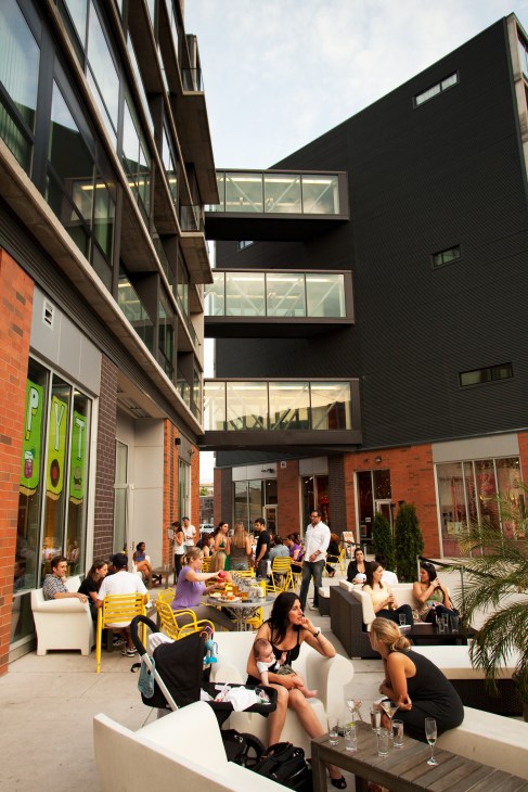 People dining and socializing in an outdoor courtyard with yellow chairs, brick buildings, and modern architectural elements.
