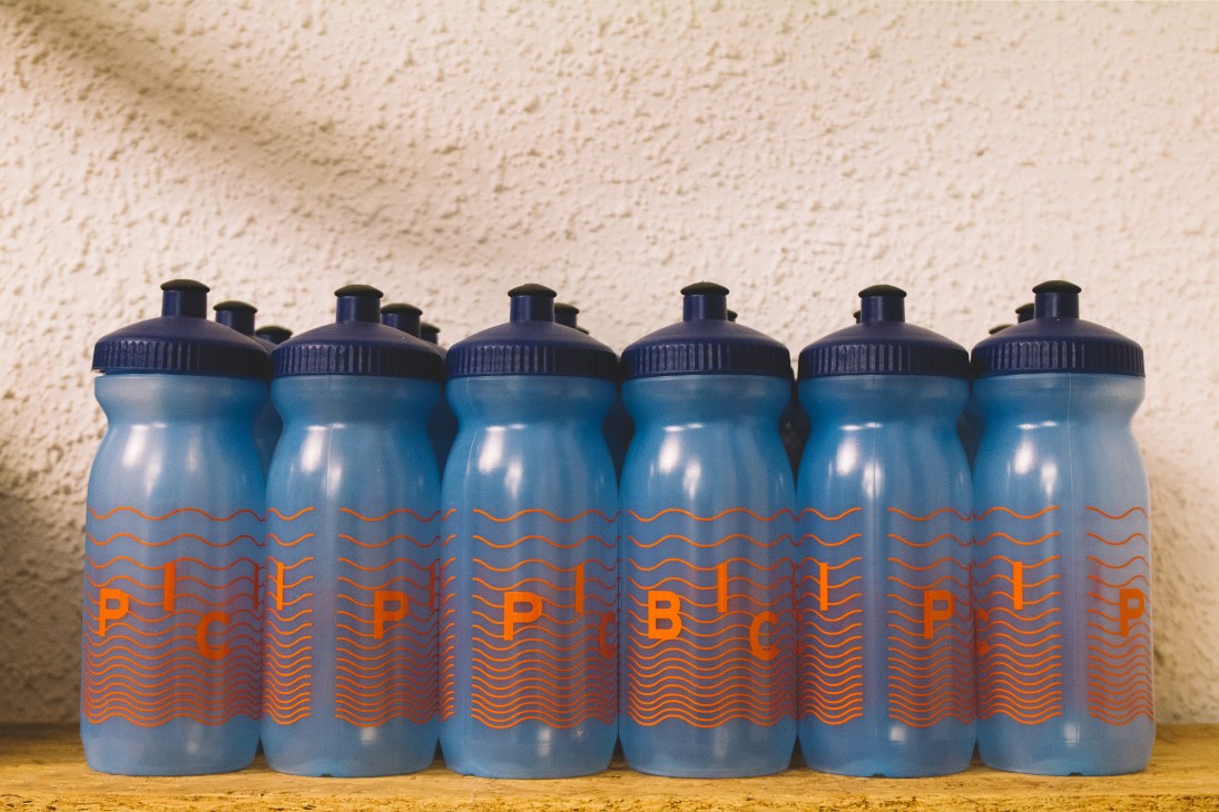 Six blue water bottles with orange wave design and 'P' branding lined up against a textured beige wall.