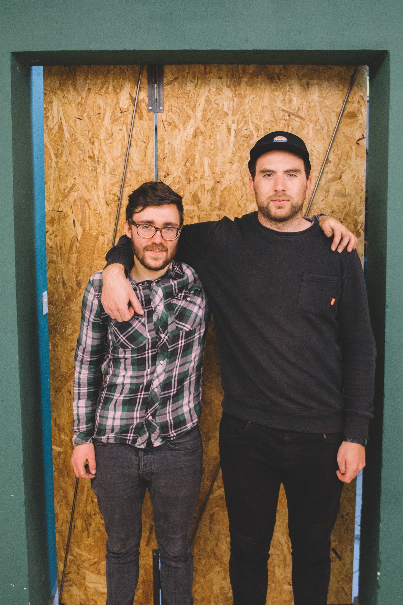 Two men pose in front of cork wall backing, one wearing glasses and plaid shirt, the other in black cap and sweatshirt.