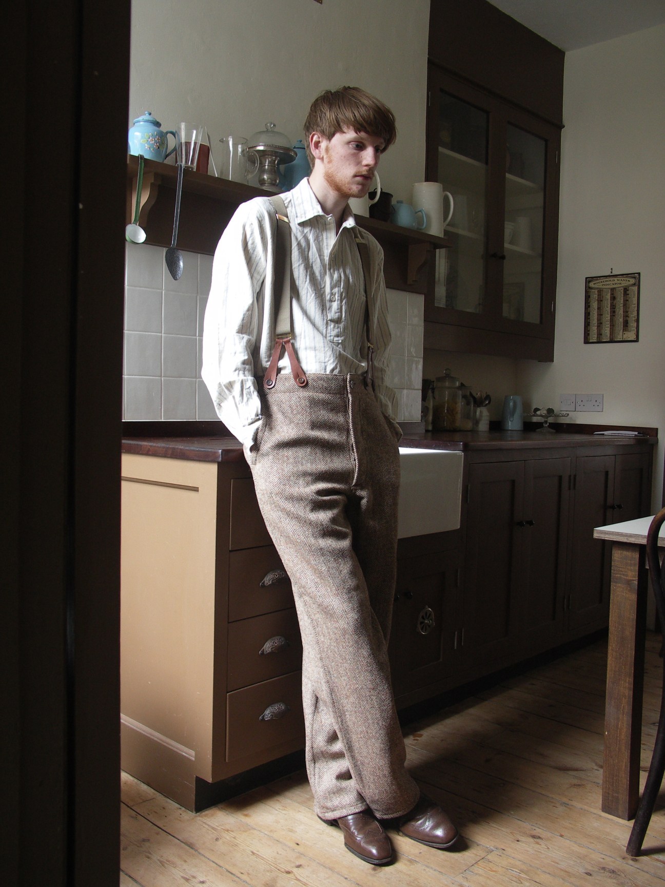 Man wearing light shirt and tweed trousers with leather suspenders in a kitchen.