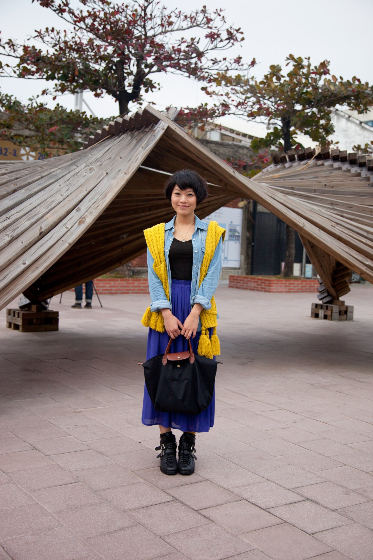 Woman in yellow vest and blue skirt standing in front of wooden sculptural structure at Pier 2, Kaohsiung.