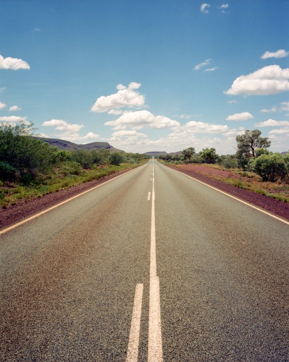 A straight asphalt road stretches toward the horizon under blue sky with white clouds, flanked by red earth and sparse green vegetation in Western Australia's outback.