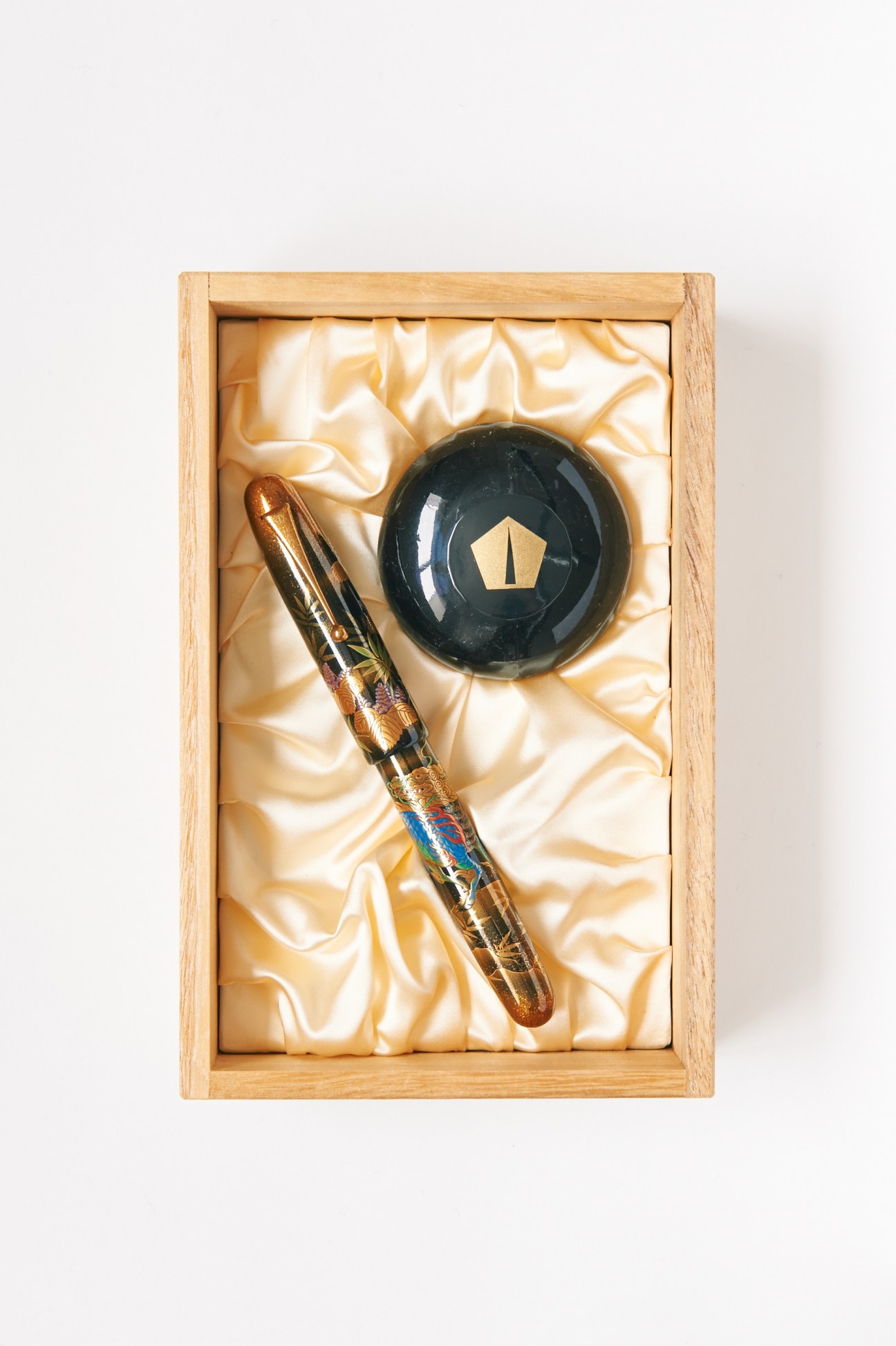 Lacquer-covered Pilot Namiki fountain pen with decorative wooden barrel next to black inkwell with gold pentagon symbol.