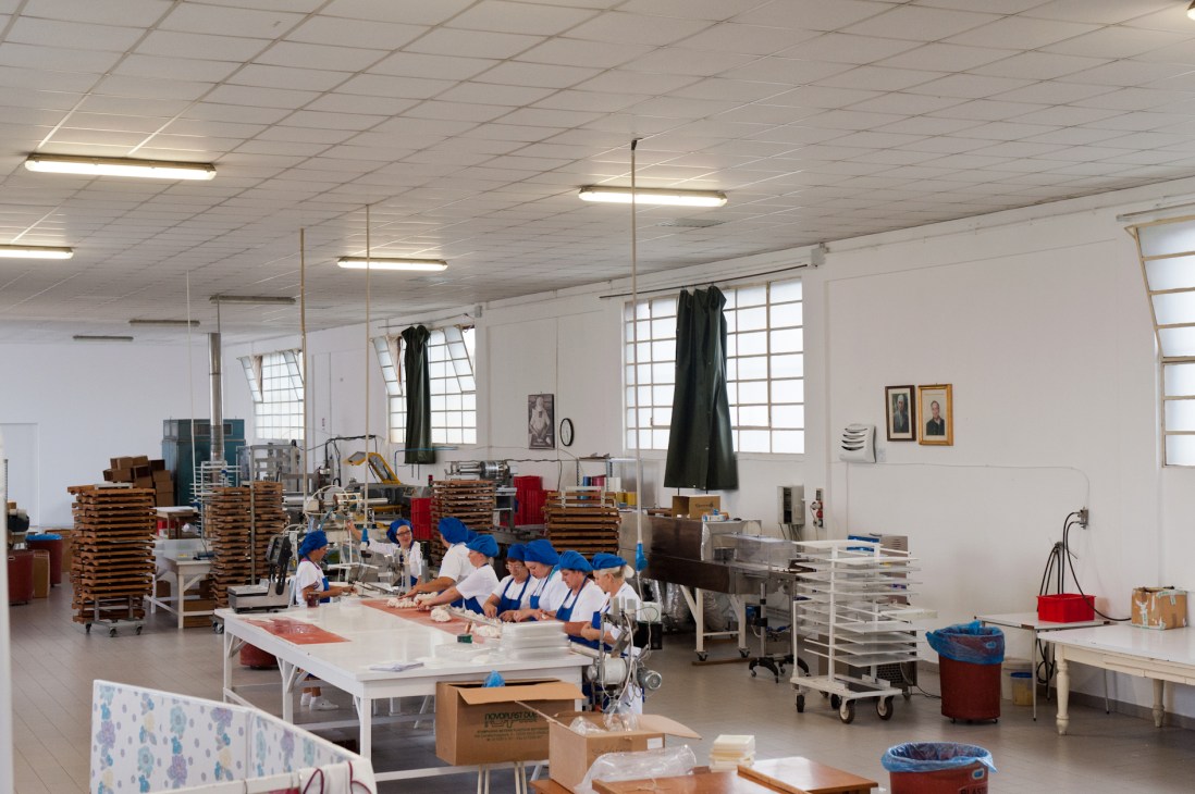 Workers in blue uniforms and caps wrap nougat at a long table in Scaldaferro's white factory laboratory.