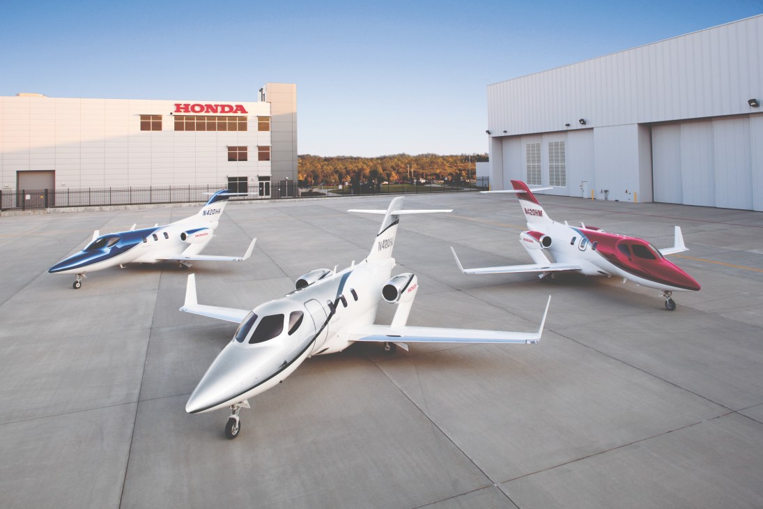 Three HondaJet business aircraft parked on tarmac in front of Honda facility with autumn forest backdrop.