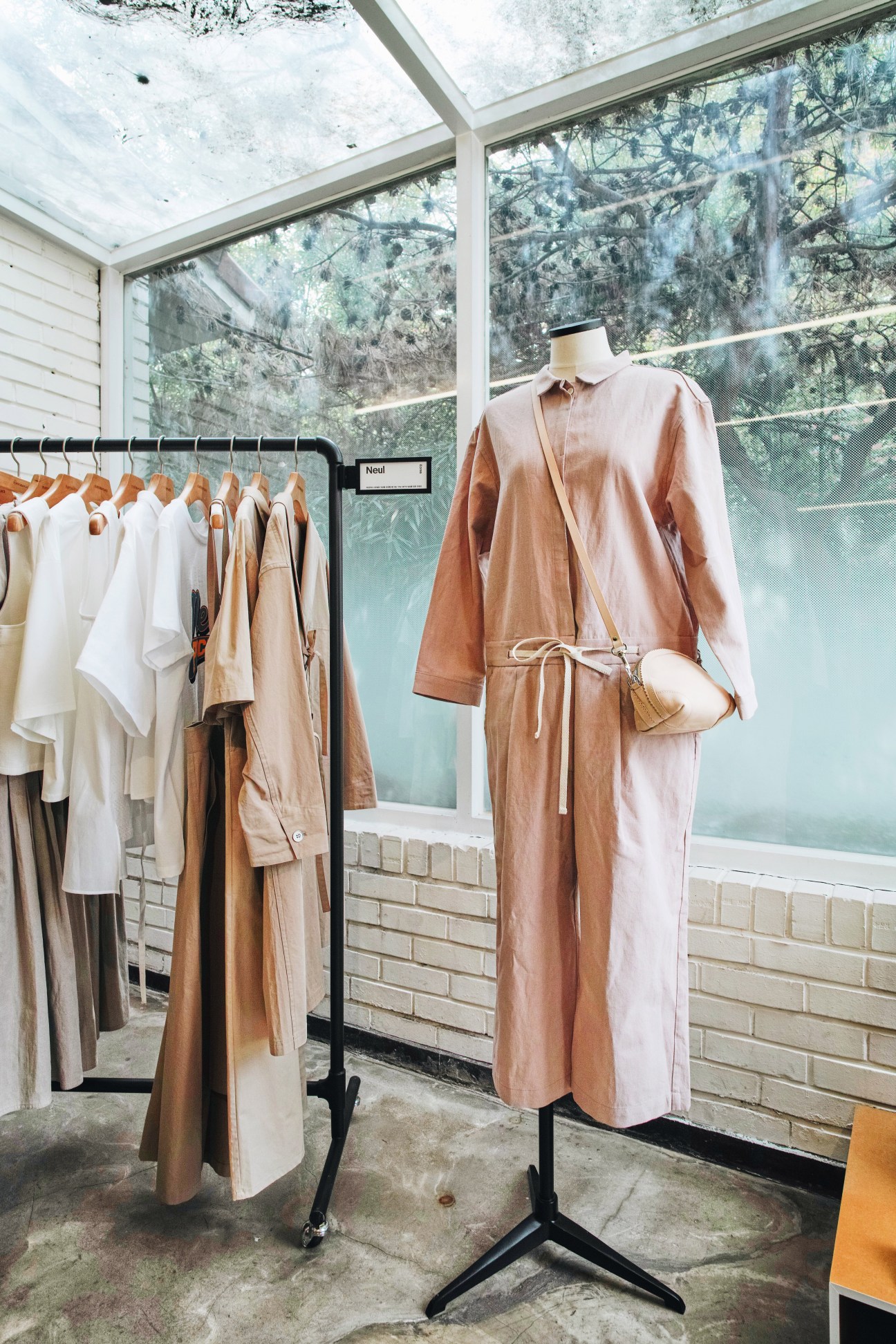 Mannequin wearing a tan linen wrap dress displayed on a black stand in a bright retail space with large windows overlooking greenery.