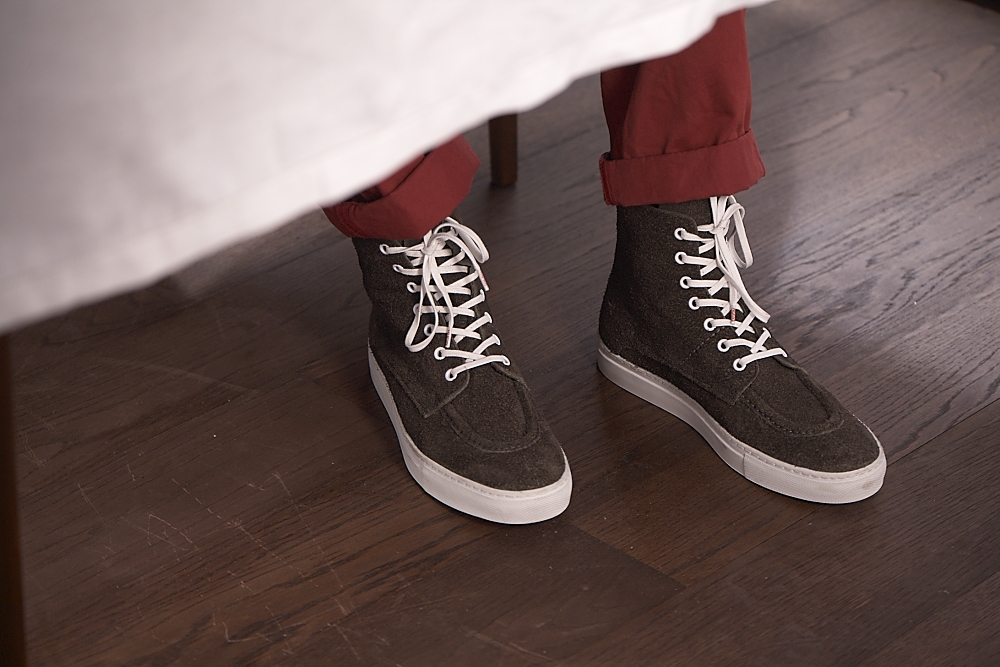 Person wearing black lace-up sneakers with white soles and burgundy rolled trousers, standing on wooden floor.