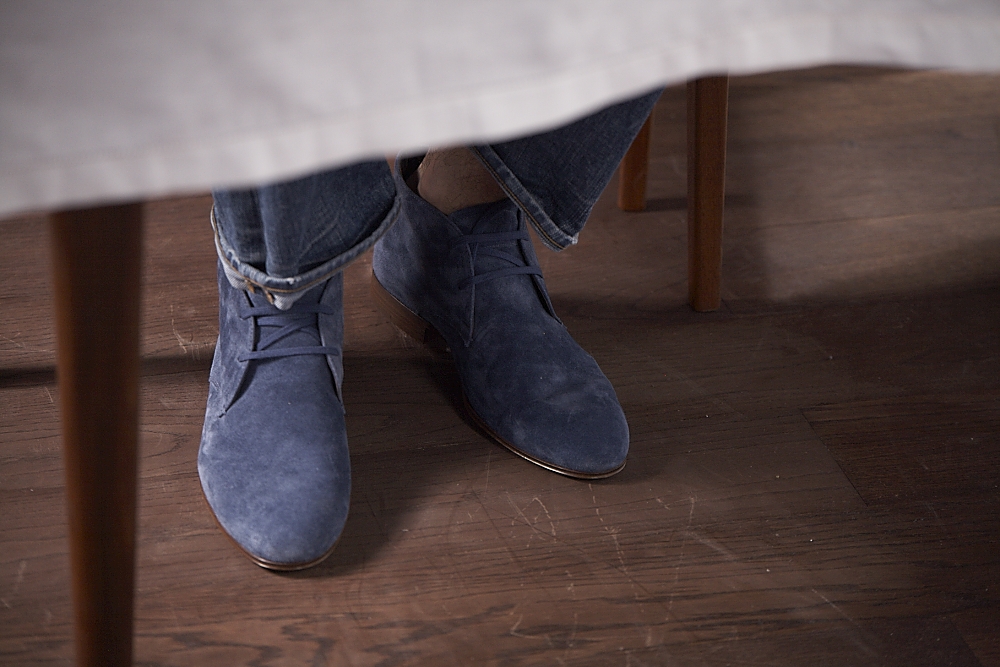 Person wearing blue suede desert boots and rolled jeans under a wooden table.
