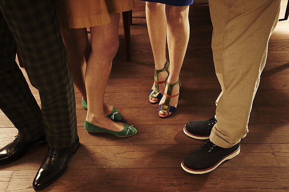 Six pairs of feet and lower legs displayed on a wooden floor, showcasing various designer shoes and clothing.