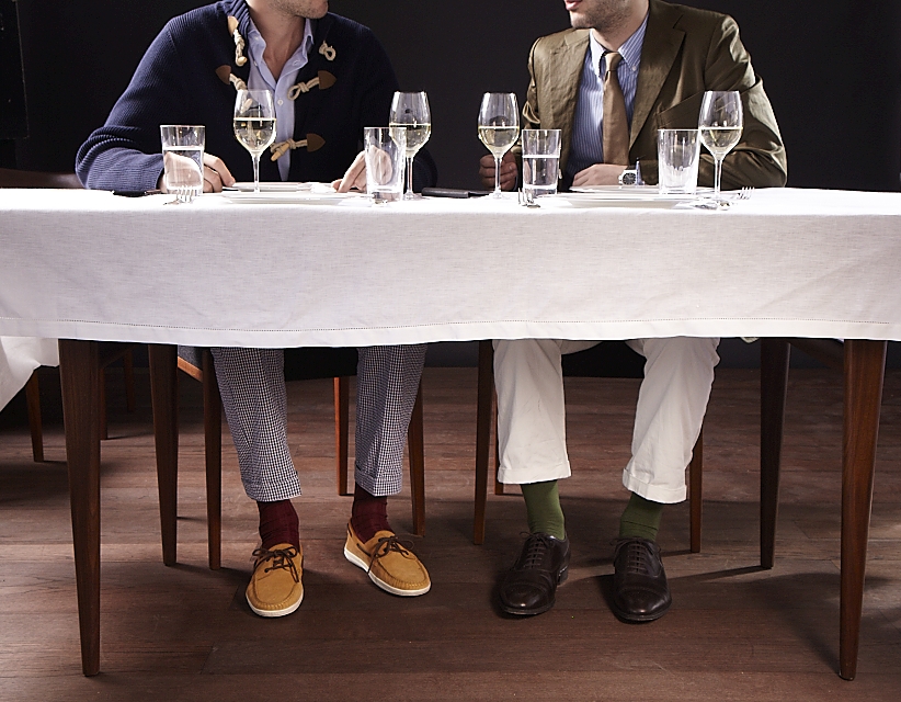 Two men seated at a dining table displaying their lower legs and footwear under a white tablecloth.