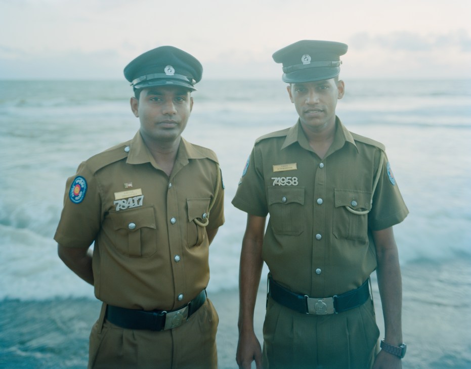 Two Sri Lankan police officers in uniforms stand together on a beach with the ocean behind them.