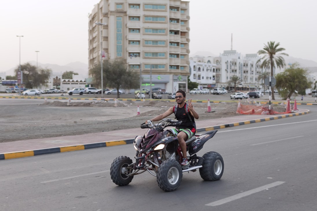 A man performs a wheelie on a three-wheeled ATV on a wide road in Fujairah with modern buildings and mountains visible in the background.