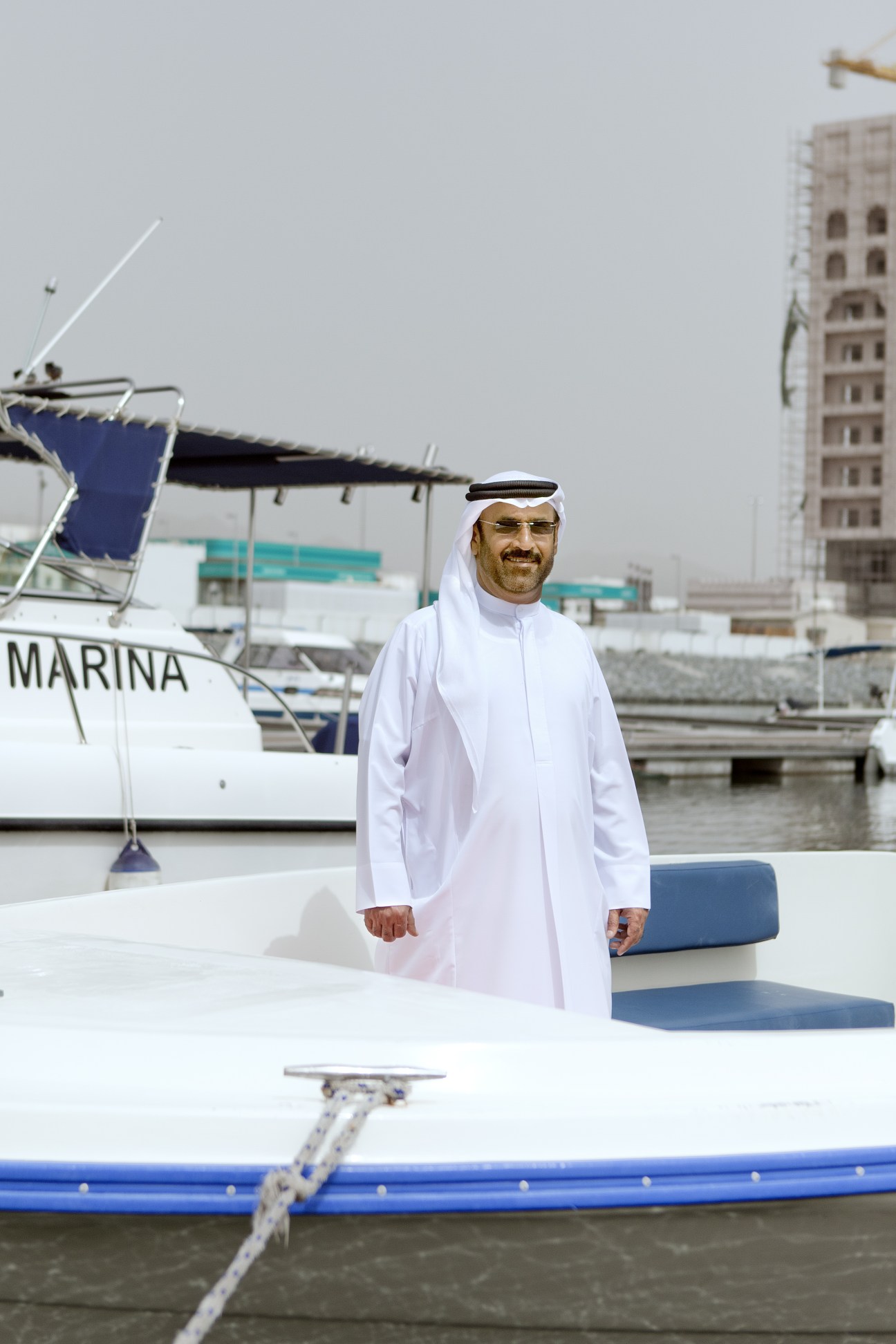 Major Ahmed Ebraheim Alblooshi stands at Fujairah International Marine Sports Club marina with boats and waterfront buildings behind him.