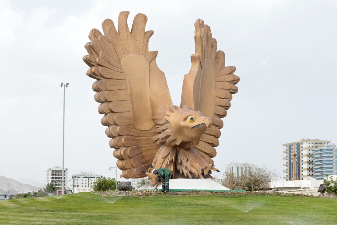 A man operates sprinklers at the base of a large wooden falcon statue on a roundabout in Fujairah, with modern buildings visible in the background.