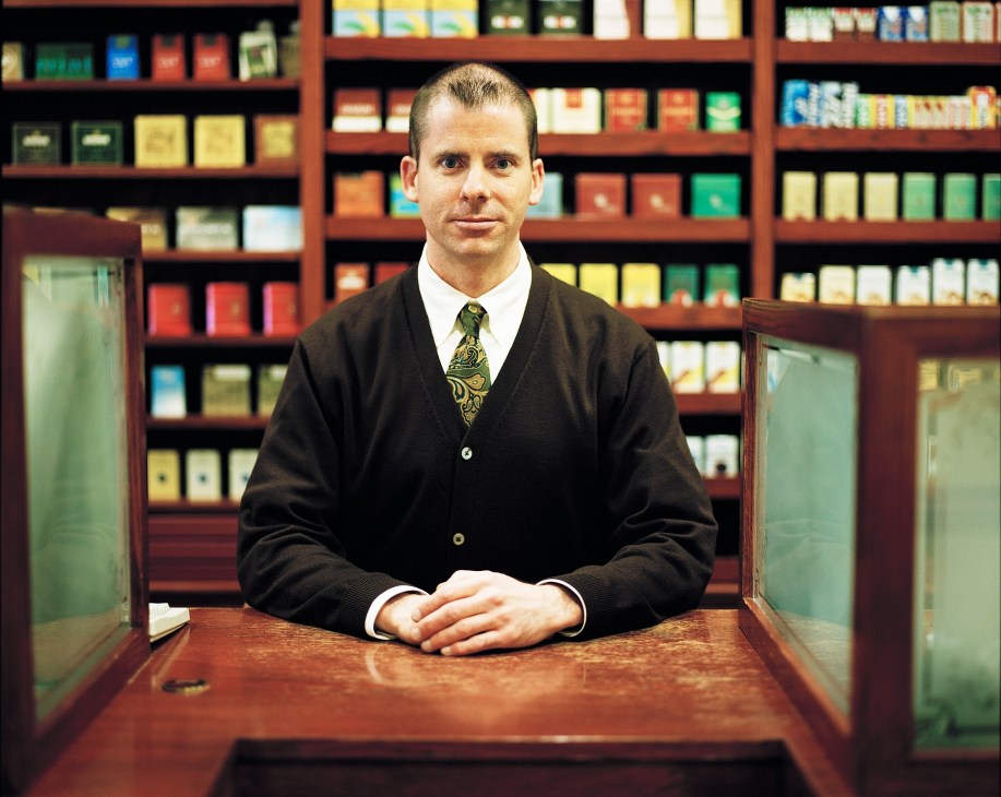Adam Smith, founder of Fog City News, stands at the counter of his San Francisco magazine shop with shelves of curated publications behind him.