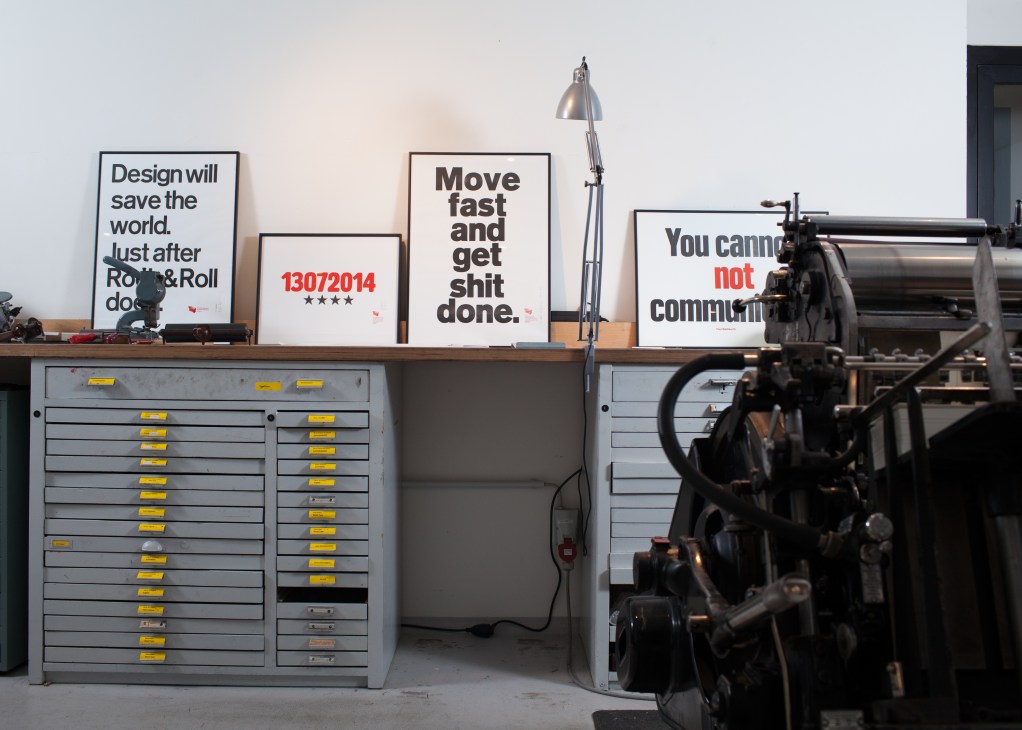 Handprinted posters with typographic messages displayed on a shelf above filing drawers and letterpress machines.