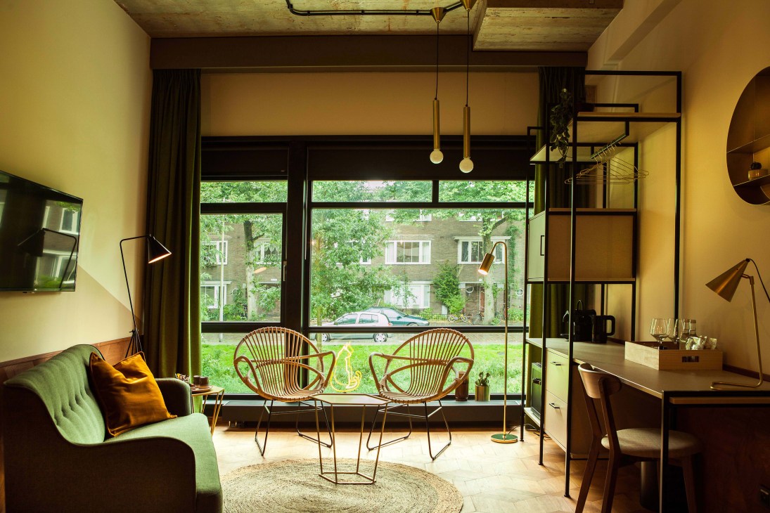 A modernist hotel room with yellow walls, green armchair, rattan chairs by large windows overlooking a street.