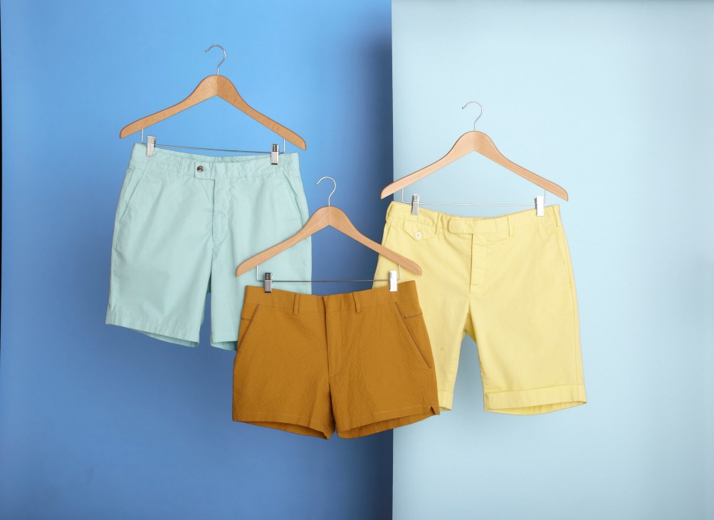 Three pairs of men's shorts in mint, mustard, and yellow hanging on wooden hangers against a blue and white background.