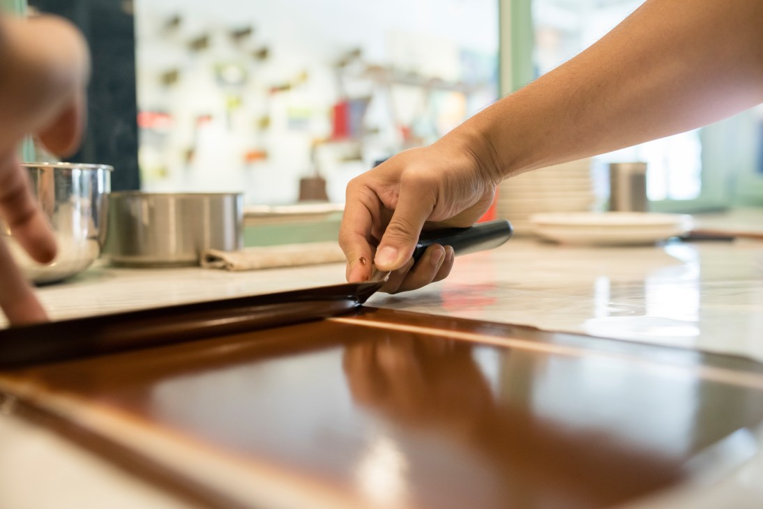 A hand holding a chocolate scraper over tempered chocolate on a work surface.