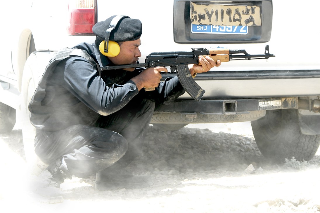 Armed security contractor in protective gear practicing tactical shooting from behind a vehicle.