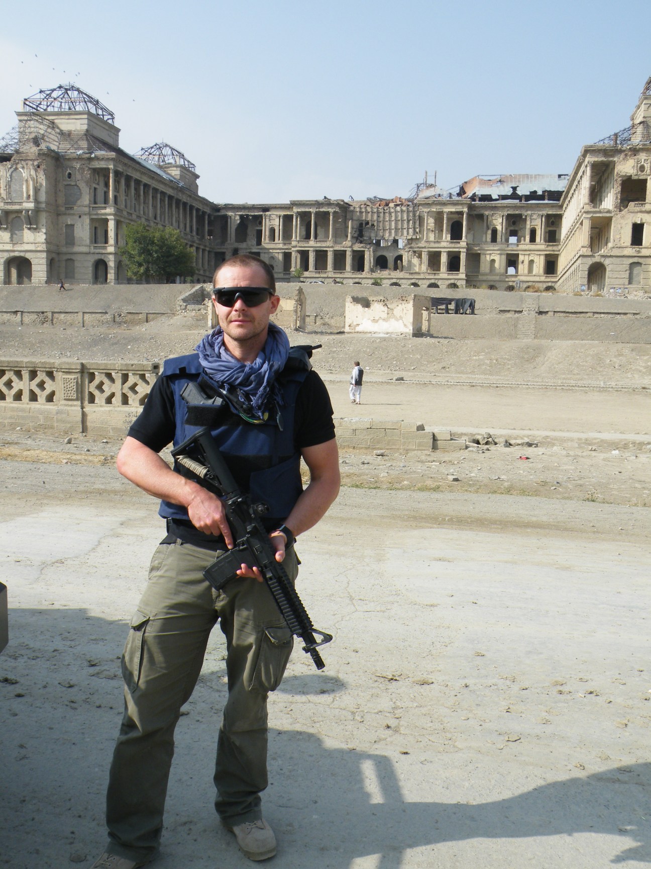 Ben Soames, founder of Fox Delta, stands armed in front of war-damaged buildings in a conflict zone.