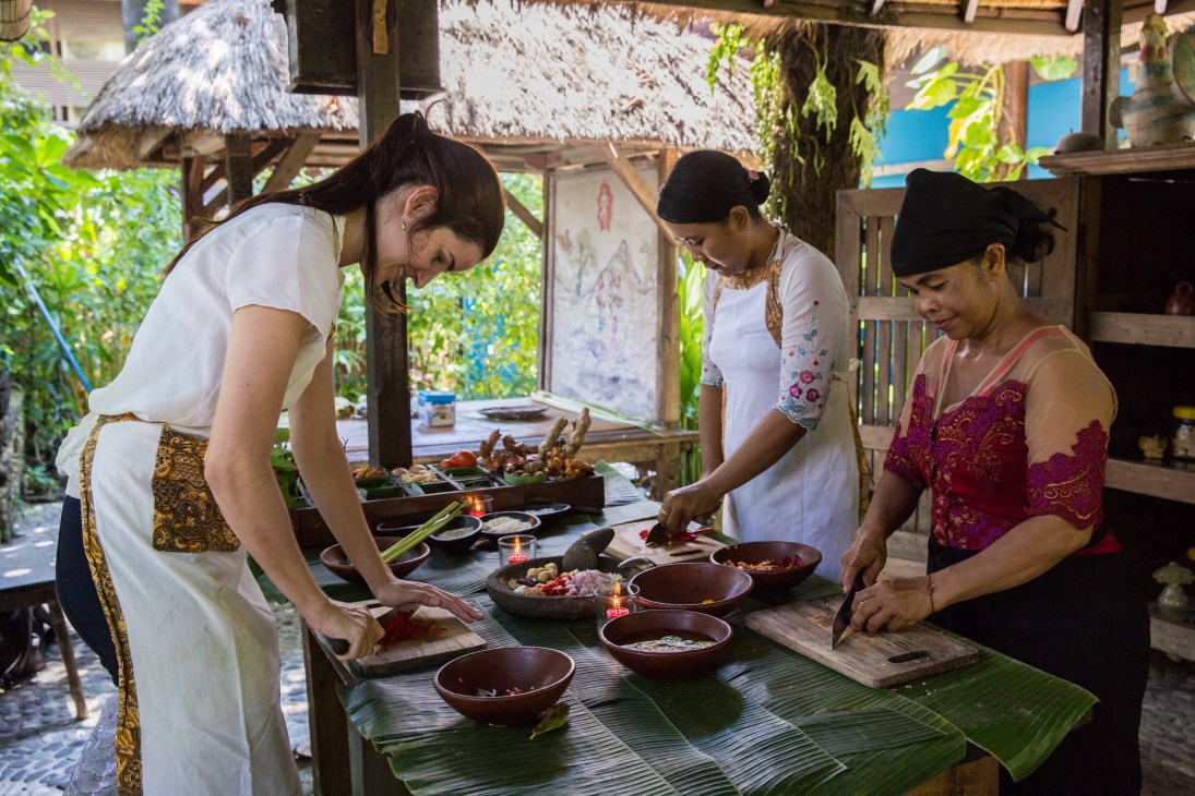 Three women prepare Balinese food together at a wooden table in a traditional open-air kitchen.