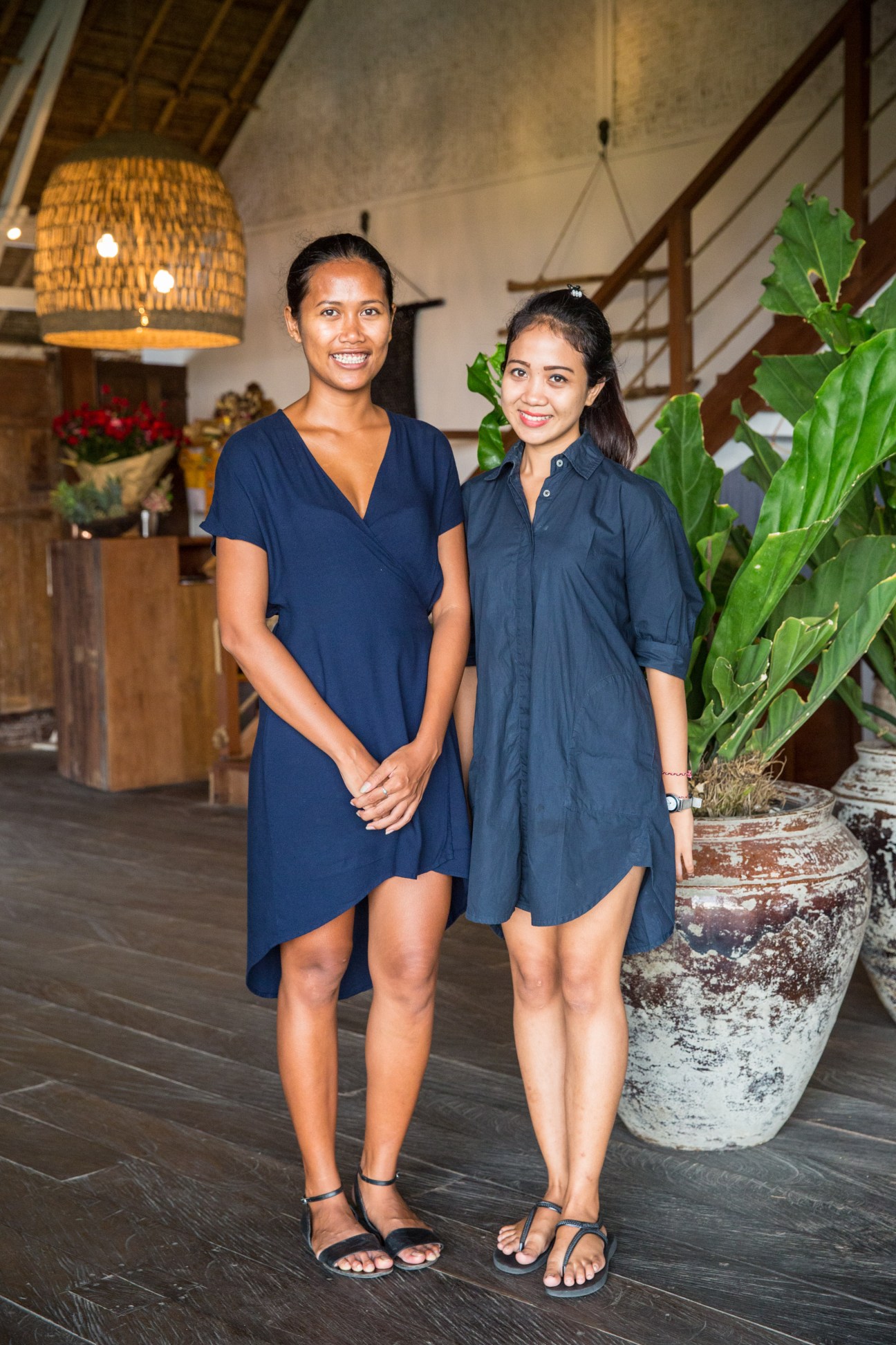 Two female staff members in navy dresses stand together in The Lawn's dining space with woven pendant lights and potted plants.