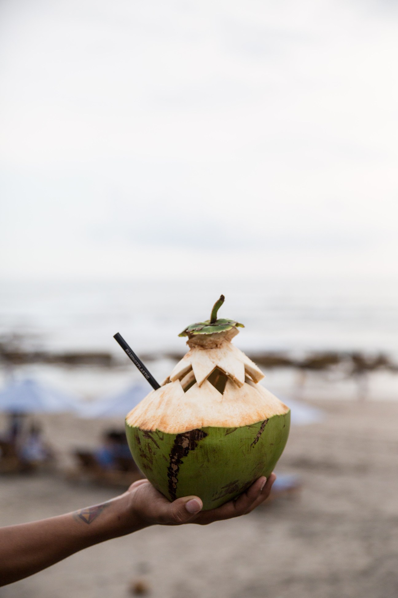 Hand holding a green coconut drink with straw and decorative coconut husk topper on a beach.