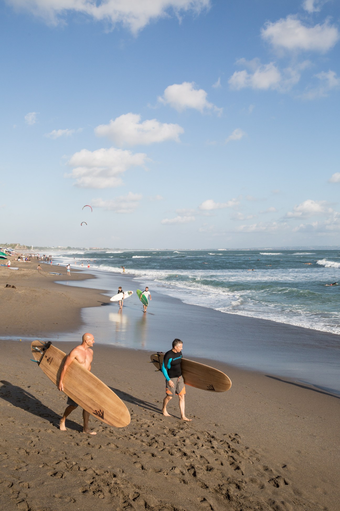 Surfers carry boards on Batu Bolong Beach with waves and kite surfers visible in the background.