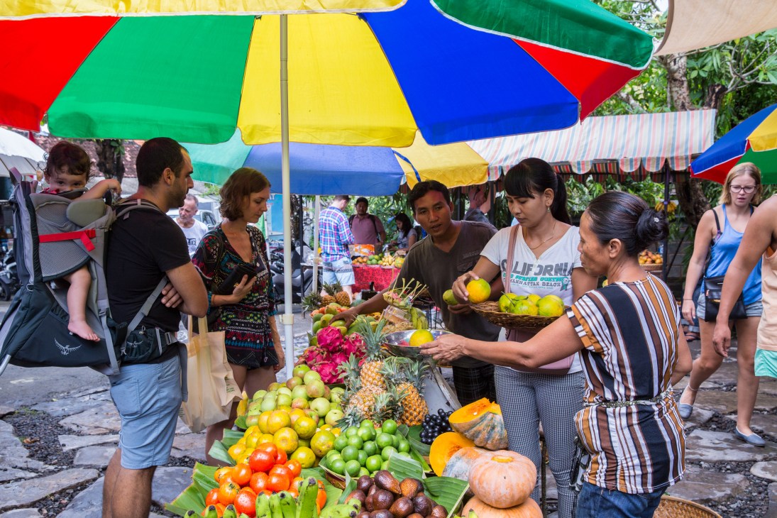 Vendors and shoppers browse fresh tropical fruits and vegetables at Samadi Sunday Organic Market under colorful umbrellas.